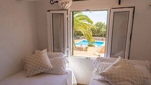 A bedroom with two beds, each with patterned pillows, and a window offering a view of a swimming pool and palm trees.