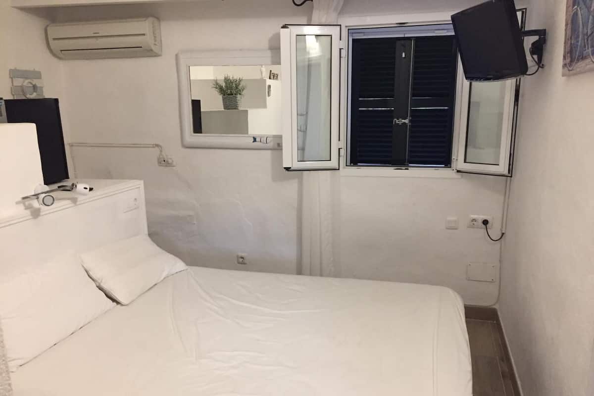 A small, white room with a bed, a window with blinds, a mirror, and a mounted TV.