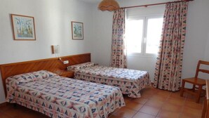 A room with two beds, a wooden headboard, a small table, a chair, and a window with curtains. - Benet Los Pinares Apartments (Santa Eulalia del Rio)