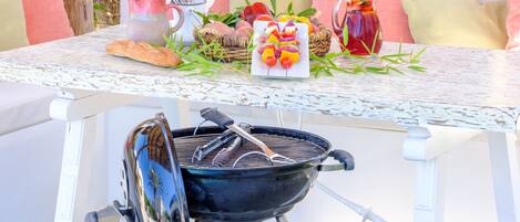 A barbecue grill on a white table with a basket of vegetables, a pitcher of drink, and a loaf of bread.