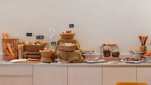 Zona de desayuno tipo buffet con panes y bollería variada, una estación de café y diferentes opciones de asientos.