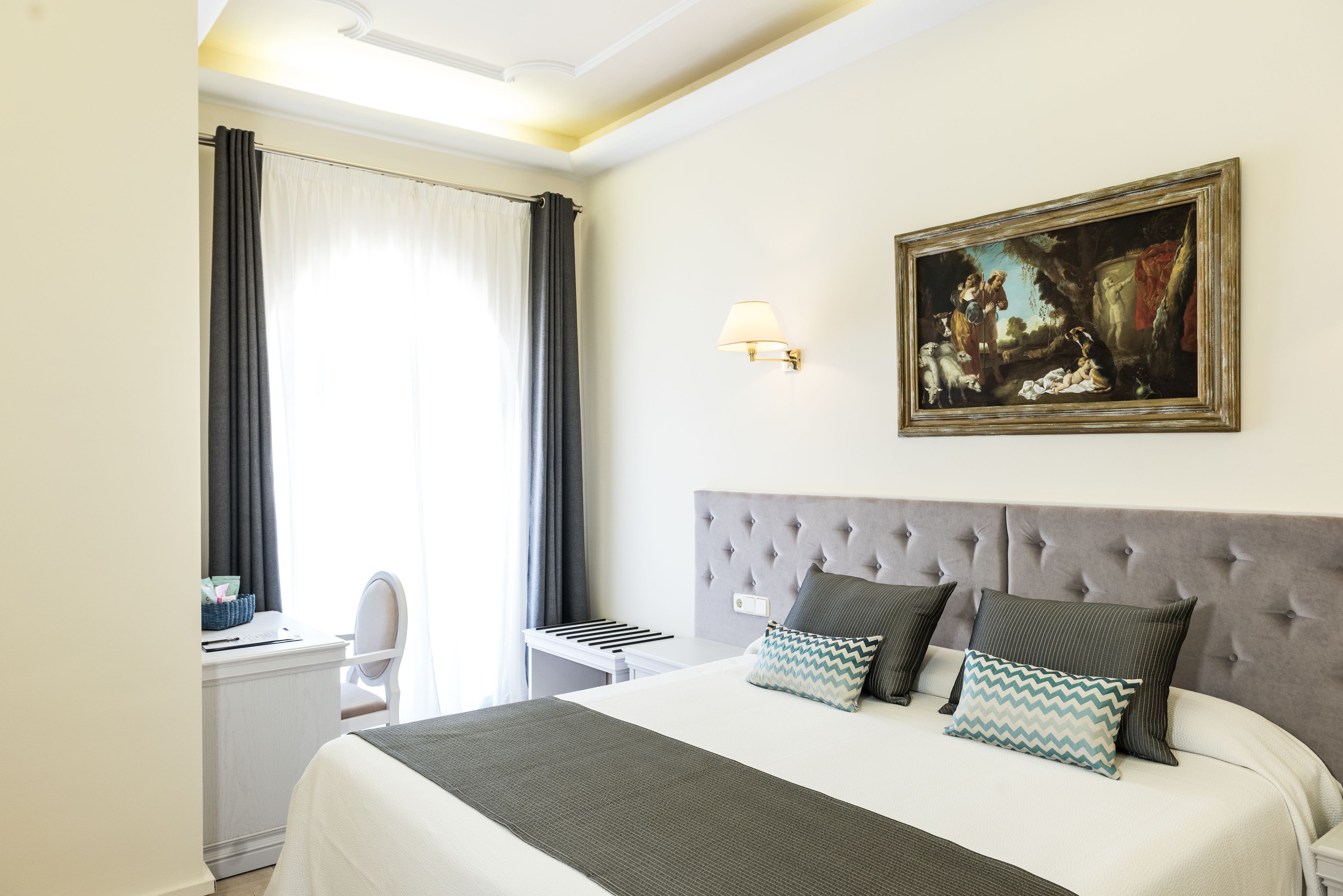 A hotel room with a large bed, a desk, and a chair. There is a painting on the wall and a lamp on the desk.