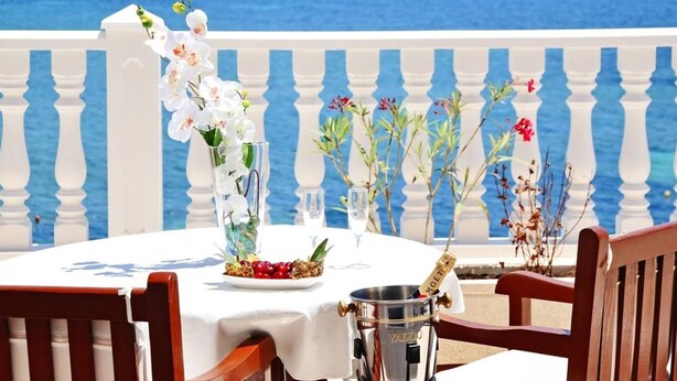 Mesa adornada con flores, una botella de champán y un plato de cerezas, con vistas al mar y una barandilla blanca.