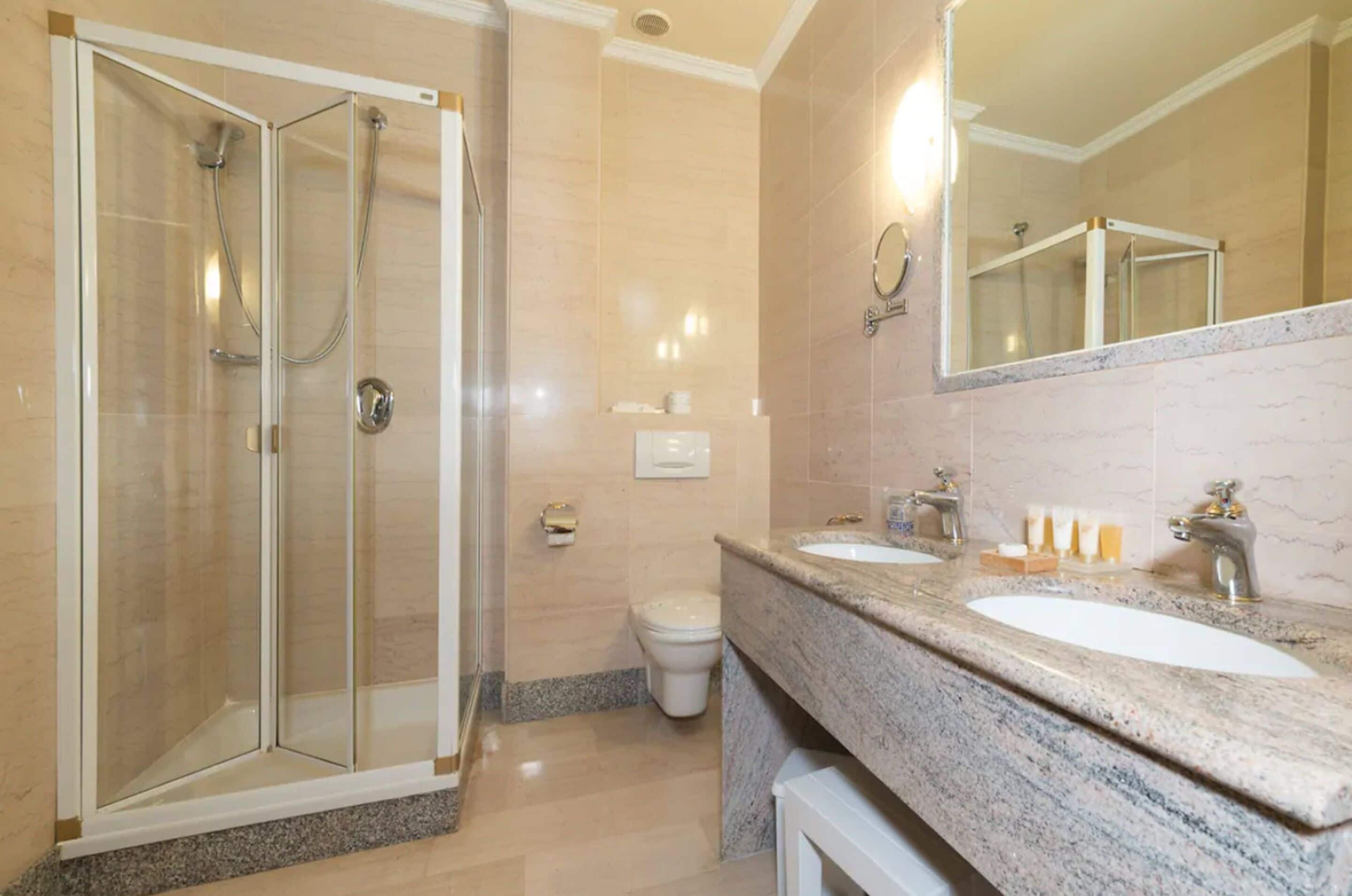 Un baño moderno con ducha de vidrio, inodoro y tocador con dos lavabos y un espejo grande.