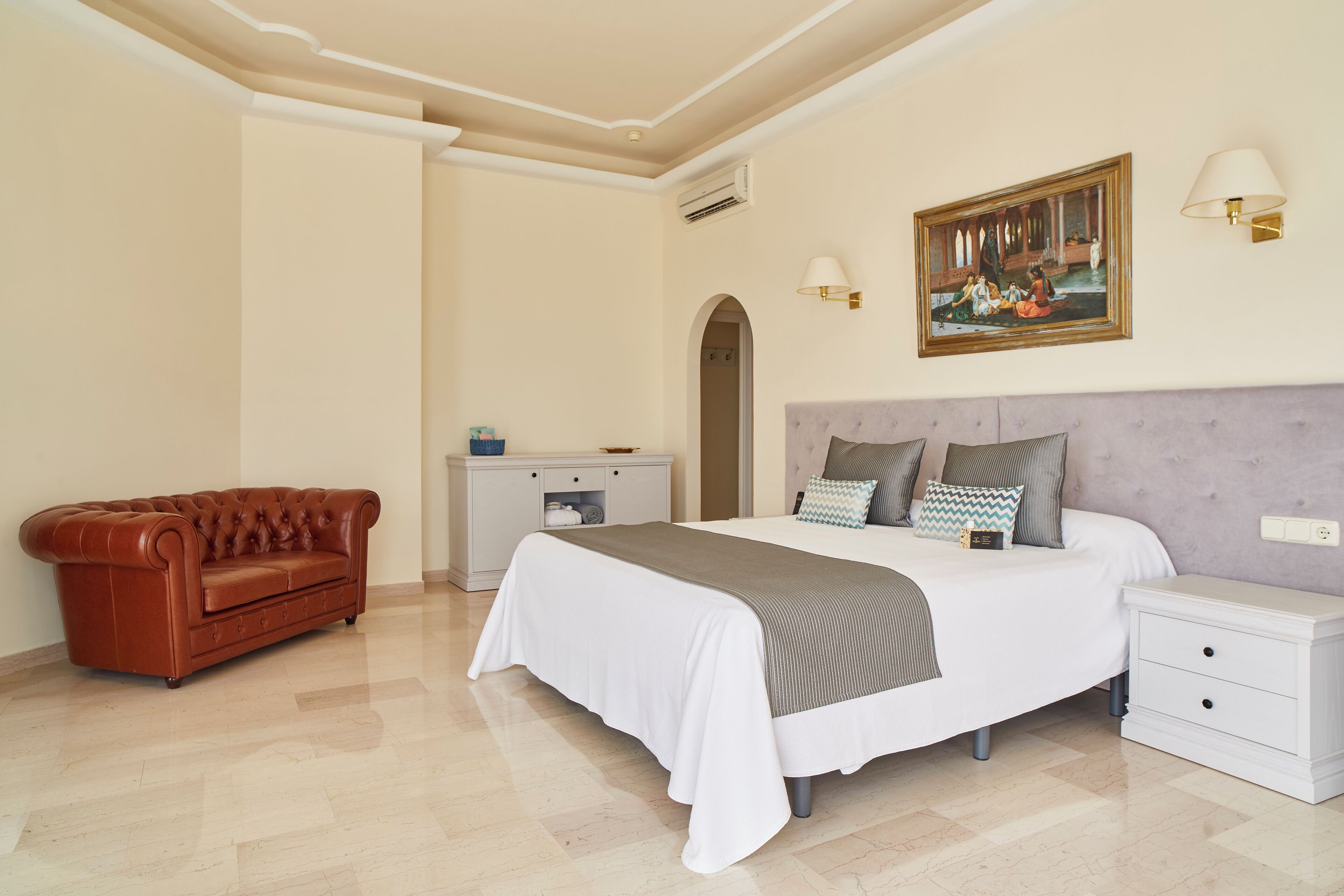 Un dormitorio con cama, mesita de noche, cómoda, sofá de cuero marrón y un cuadro en la pared.