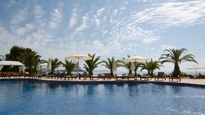 Zona junto a la piscina con sillones de descanso y sombrillas, palmeras y cielo despejado.