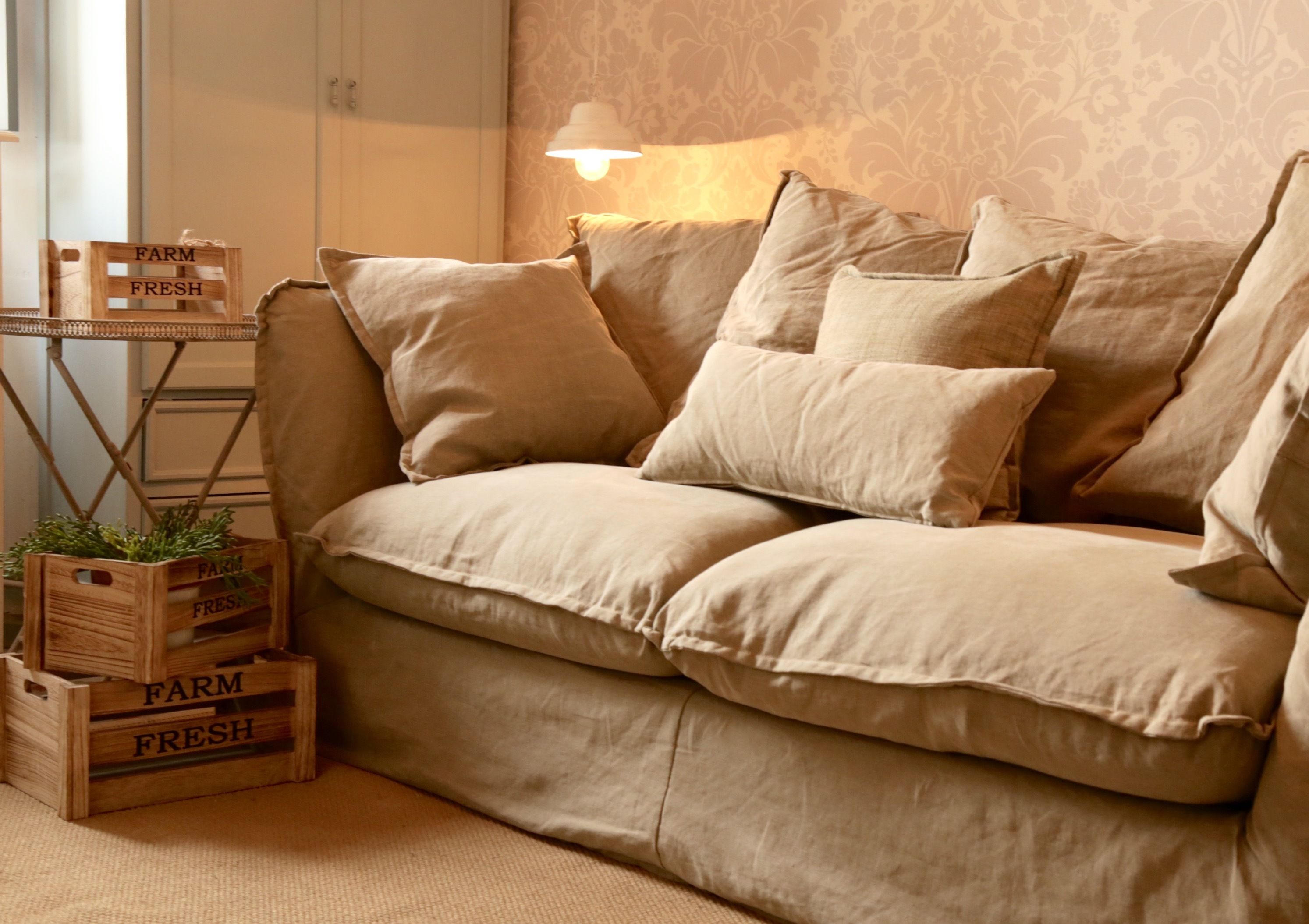 Een beige bank met meerdere kussens, een houten kist met de tekst 'FARM FRESH' en planten, en een wandlamp.