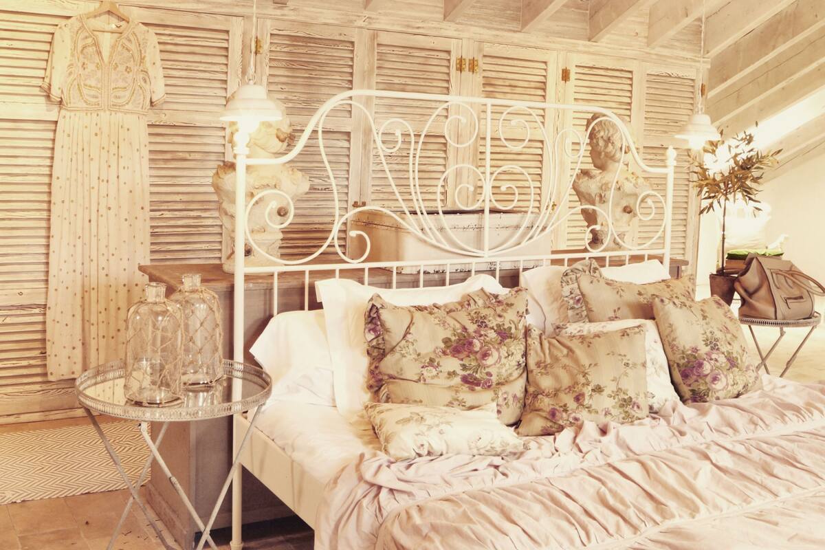 Ein Schlafzimmer mit weißem Metallbettgestell, floralen Kissen, einer Hängelampe und einem kleinen runden Tisch mit Glasflaschen.