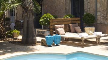 A poolside area with a wooden bench, cushions, and potted plants.