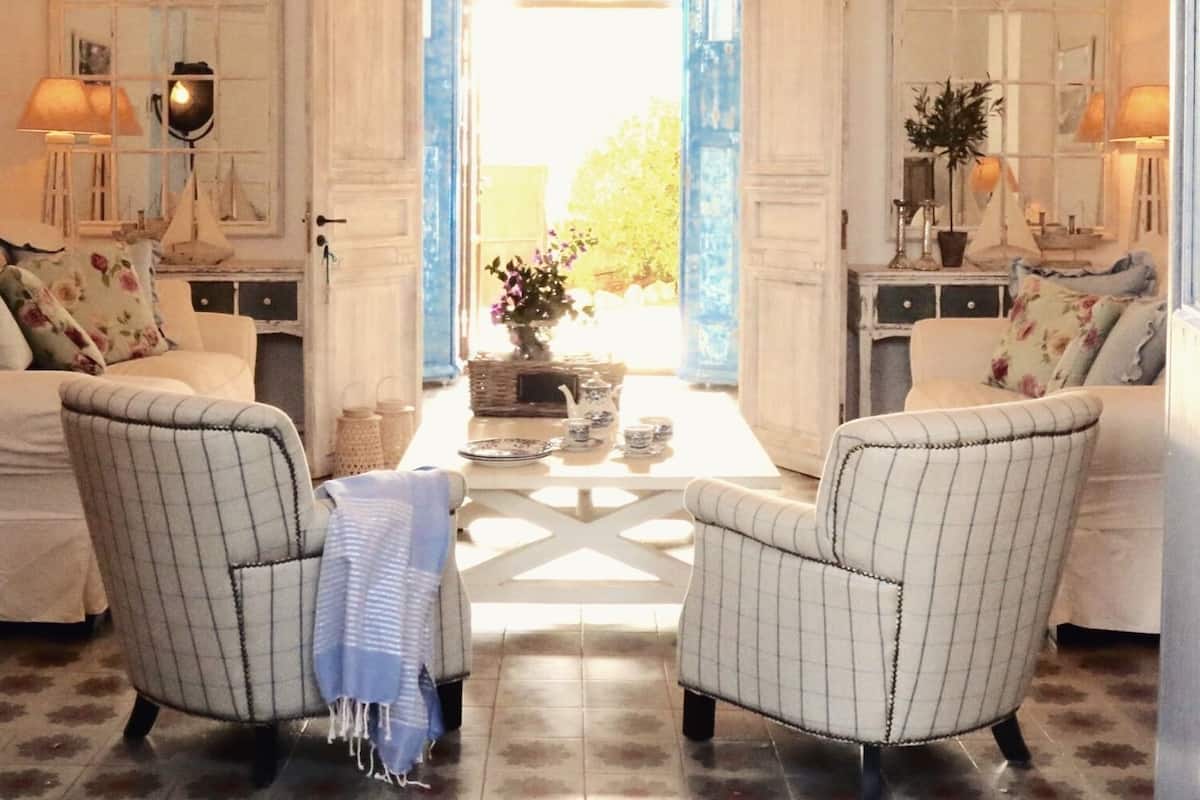Ein Zimmer mit zwei Polstersesseln, die eine Tisch mit Blumen, einer Chandelieber und Blick durch eine offene Tür zeigen.