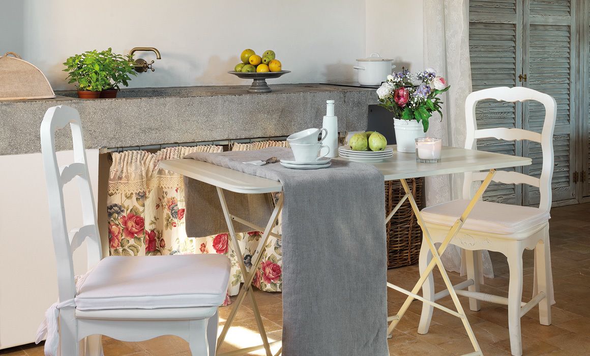 Un comedor con una mesa dispuesta para dos personas, una silla blanca, un banco, una planta en maceta, un cuenco de fruta y un florero con flores.