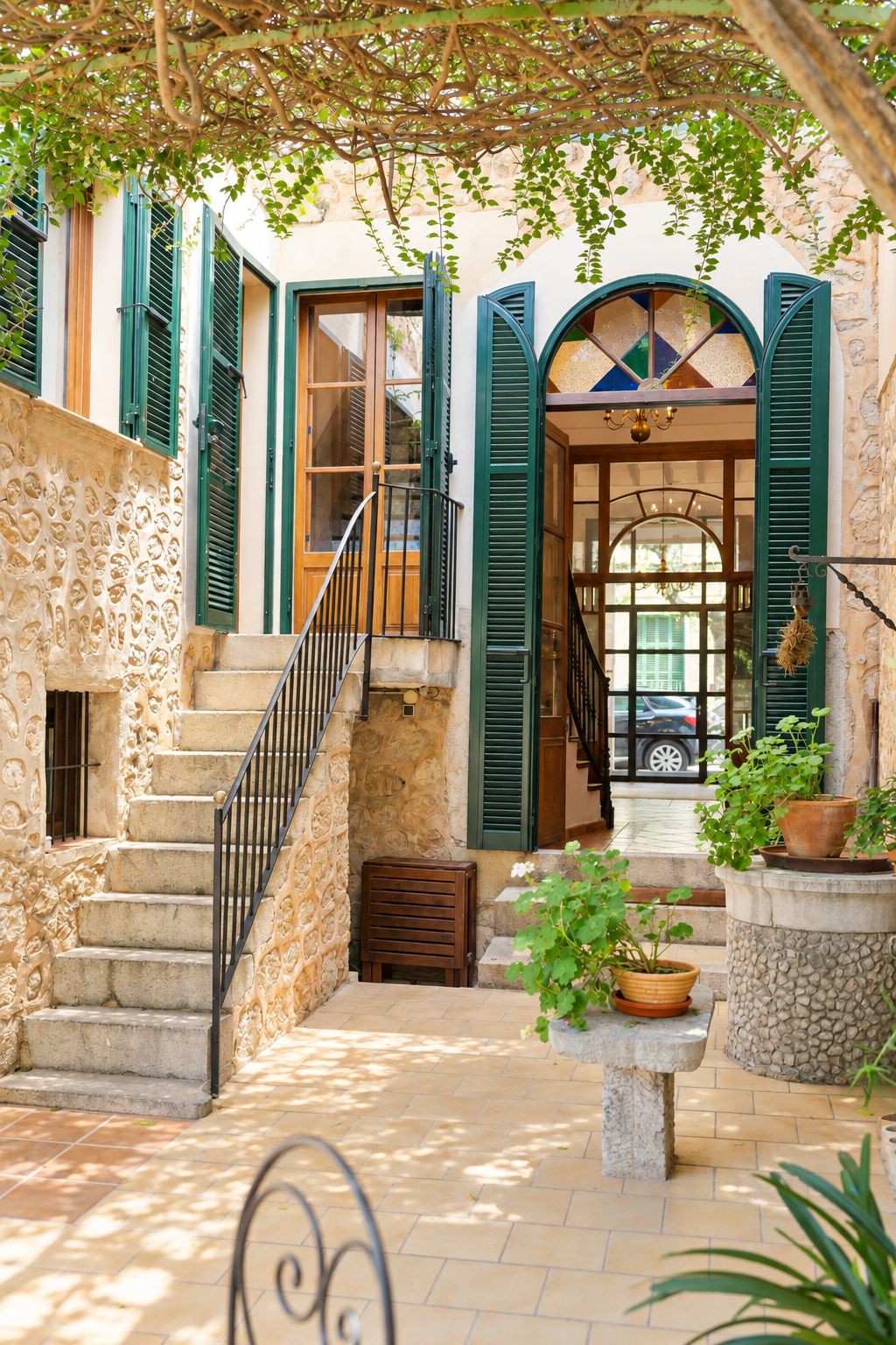 Una escalera de piedra que lleva a una puerta de madera con contraventanas verdes, un banco pequeño y plantas en macetas.