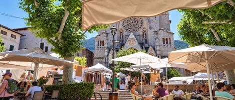 An outdoor café area with tables and chairs under umbrellas, a stone building with a clock tower in the background, and people dining and walking around.