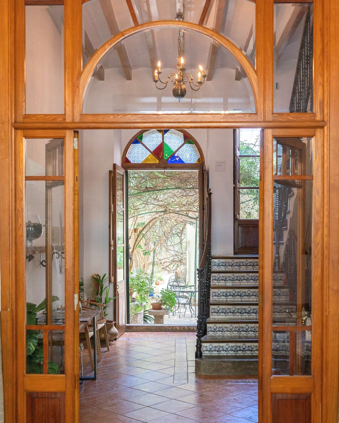 Un passage surmonté d’un cadre en bois, donnant sur un patio carrelé, avec un escalier et une fenêtre à vitraux.