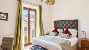 A bedroom with a large bed, a chair, a nightstand, a lamp, and a view of the outside.