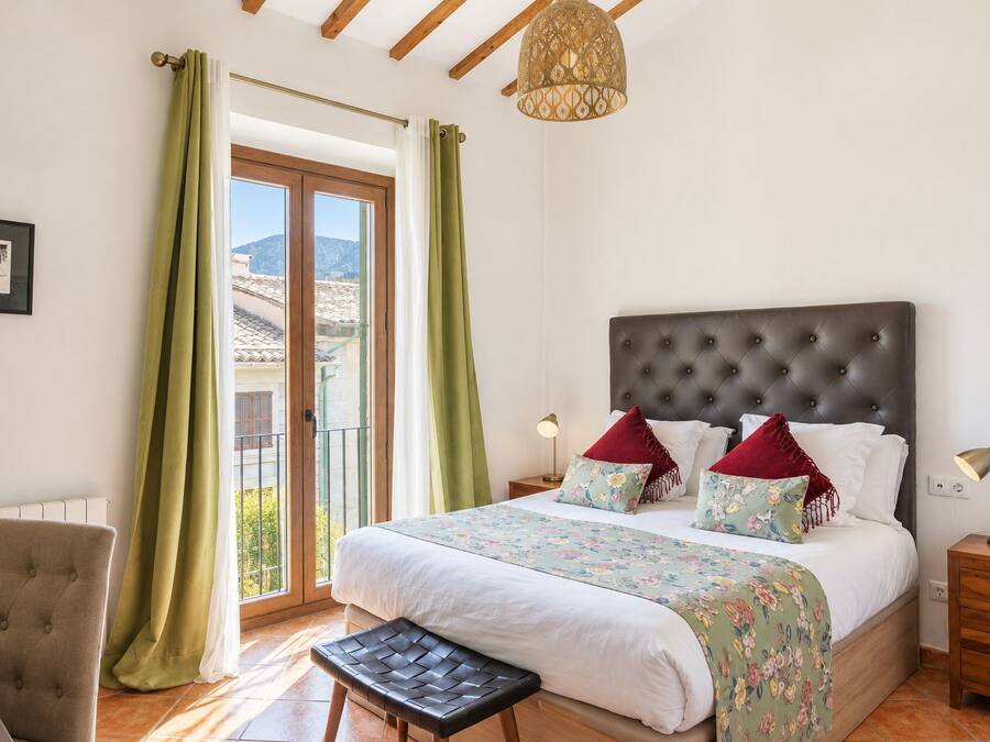 A bedroom with a large bed, a chair, a nightstand, a lamp, and a view of the outside.