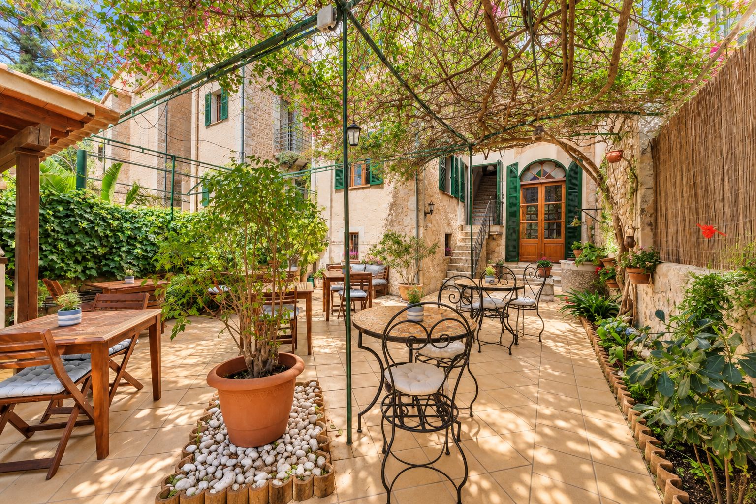 Une terrasse extérieure avec des tables et des chaises en bois, entourée de plantes en pot et d’un mur de pierre.