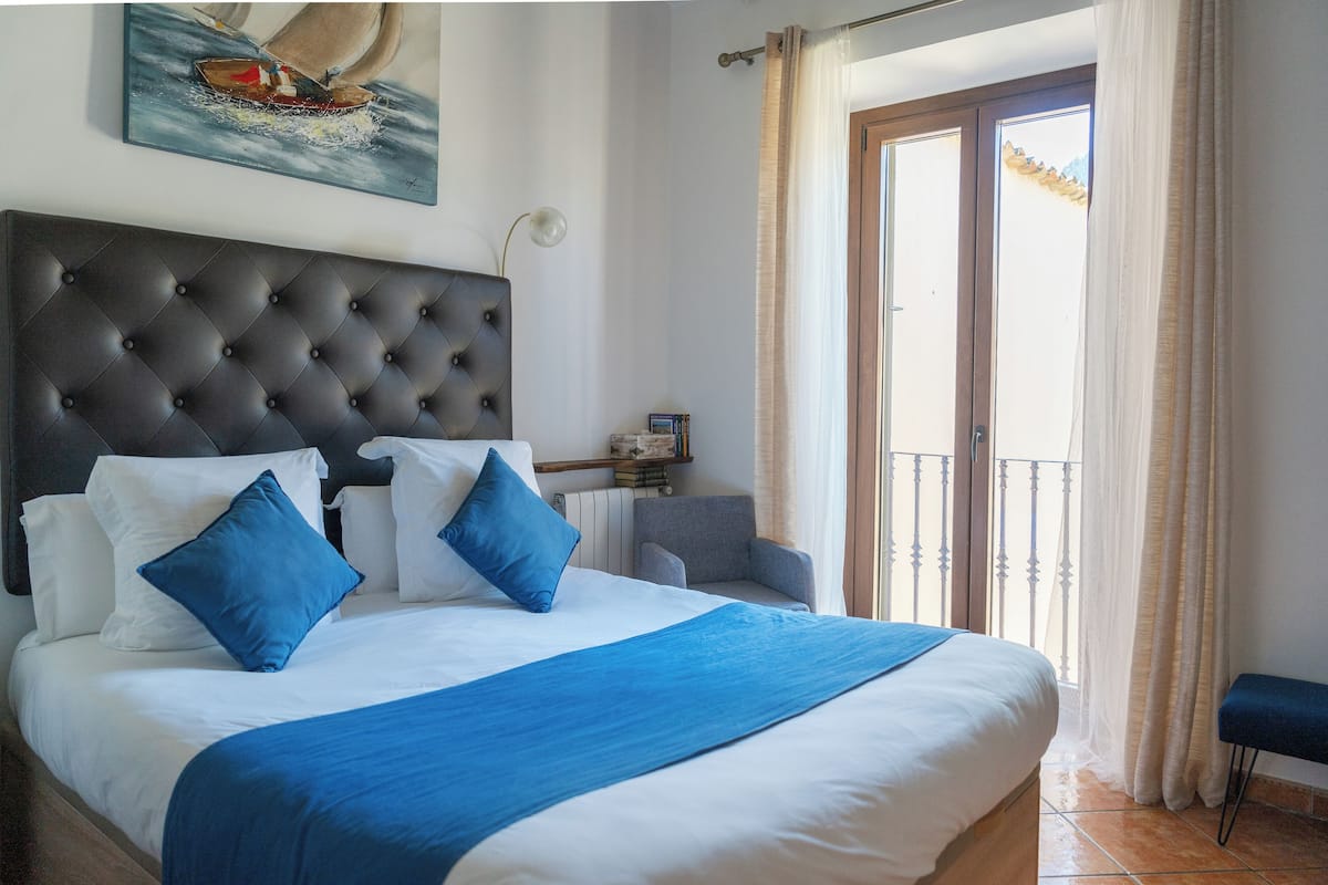 Un lit bien fait, doté d’une tête de lit capitonnée, d’un plaid bleu et de coussins assortis. On aperçoit une petite table d’appoint avec une lampe et des livres.