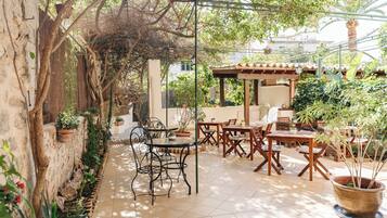 An outdoor patio area with a pergola, wooden tables and chairs, potted plants, and a stone wall.