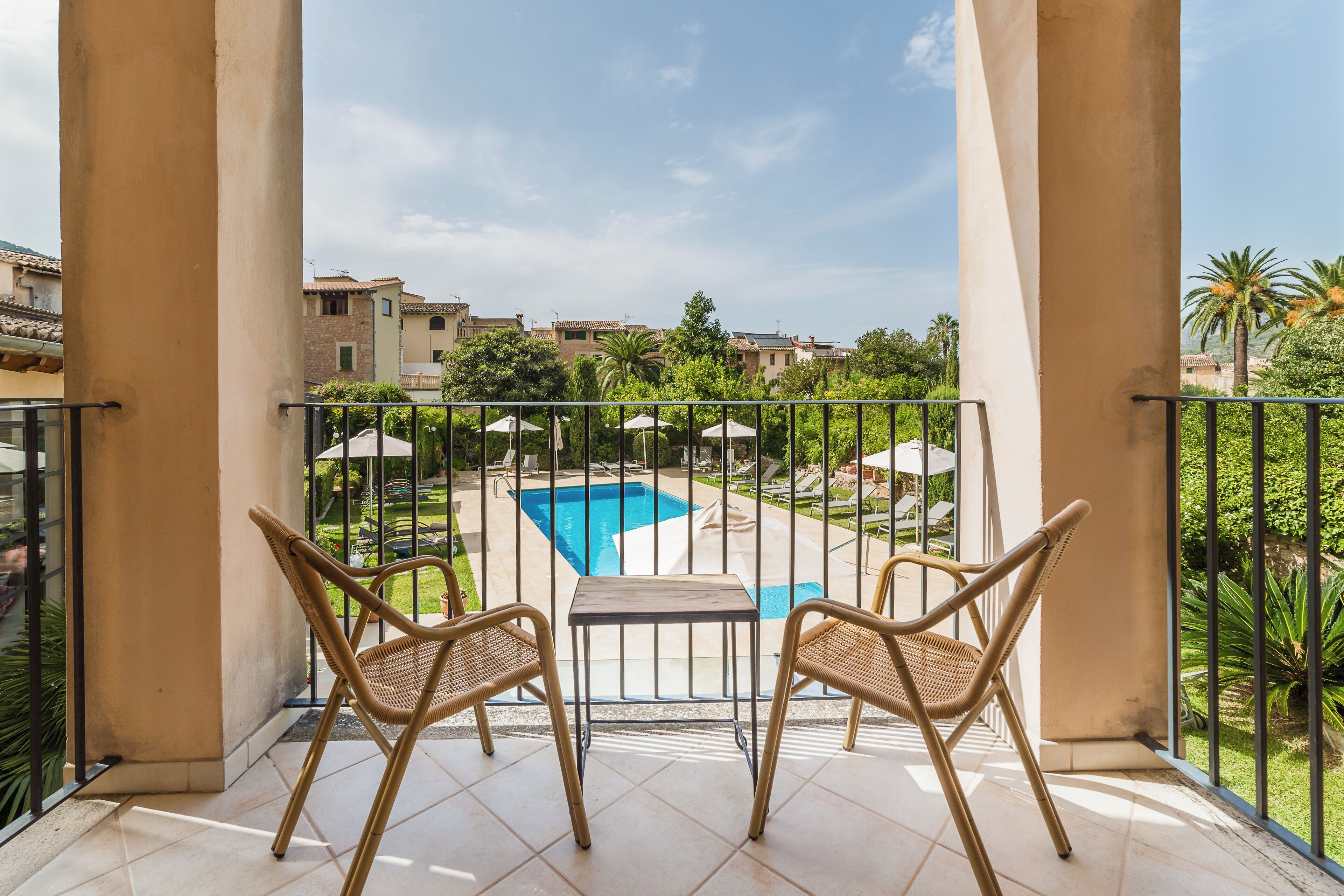 Un balcón con dos sillas de mimbre y una mesita, con vista a una piscina y abundante vegetación.