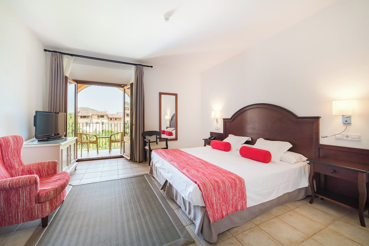 A hotel room with a bed, a desk, a chair, a TV, and a balcony with a view of buildings.