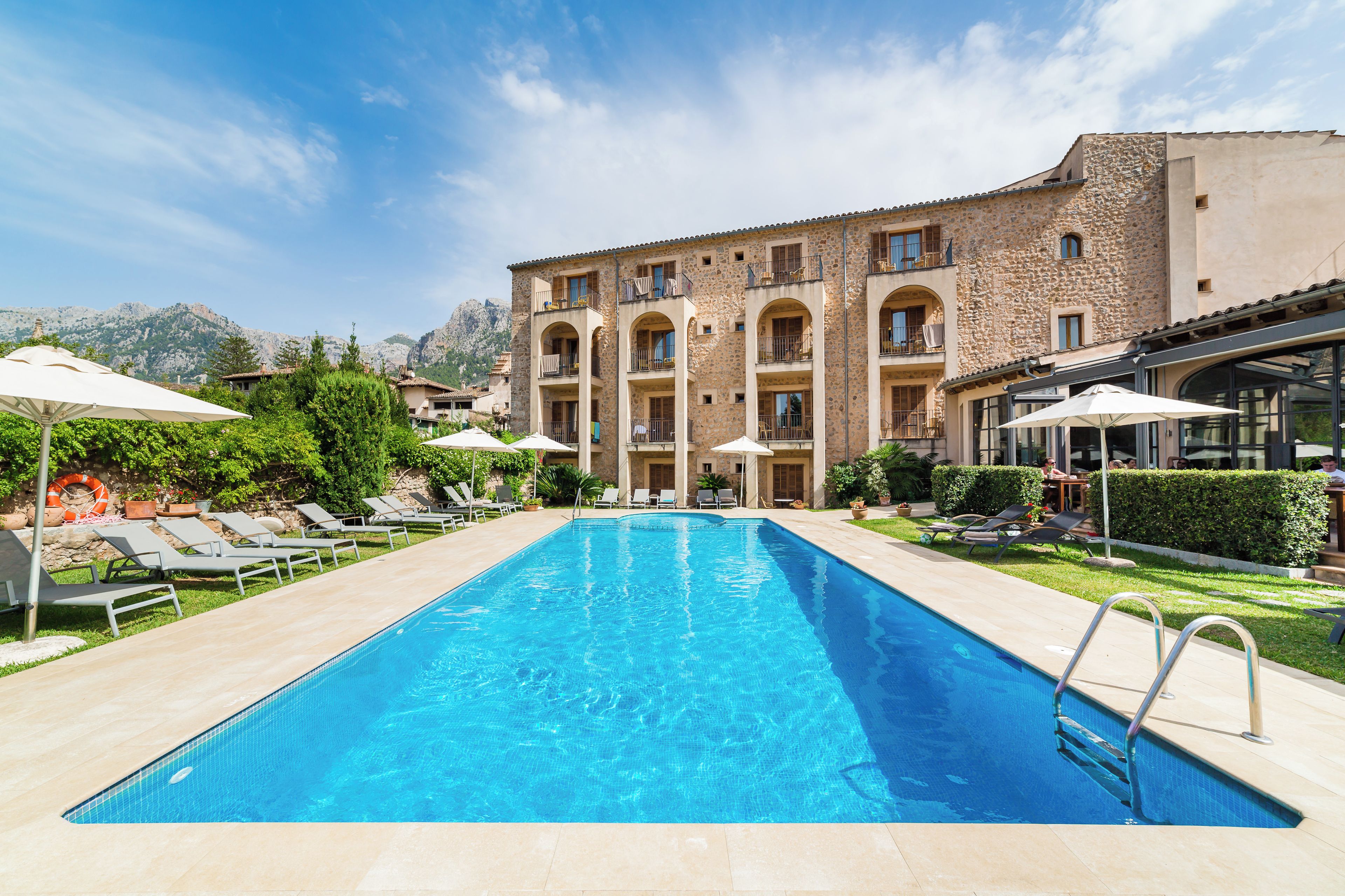 A swimming pool with lounge chairs and umbrellas, surrounded by a building with multiple balconies and a mountainous backdrop.