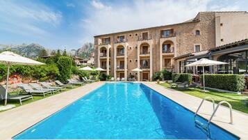 A swimming pool with lounge chairs and umbrellas, surrounded by a building with multiple balconies and a mountainous backdrop.