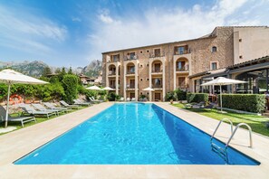 A swimming pool with lounge chairs and umbrellas, surrounded by a building with multiple balconies and a mountainous backdrop. - Ca'l Bisbe (Sóller)