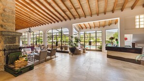 Un vaste espace intérieur doté d’une cheminée en pierre, de poutres apparentes en bois, de grandes fenêtres et offrant une vue sur la piscine.