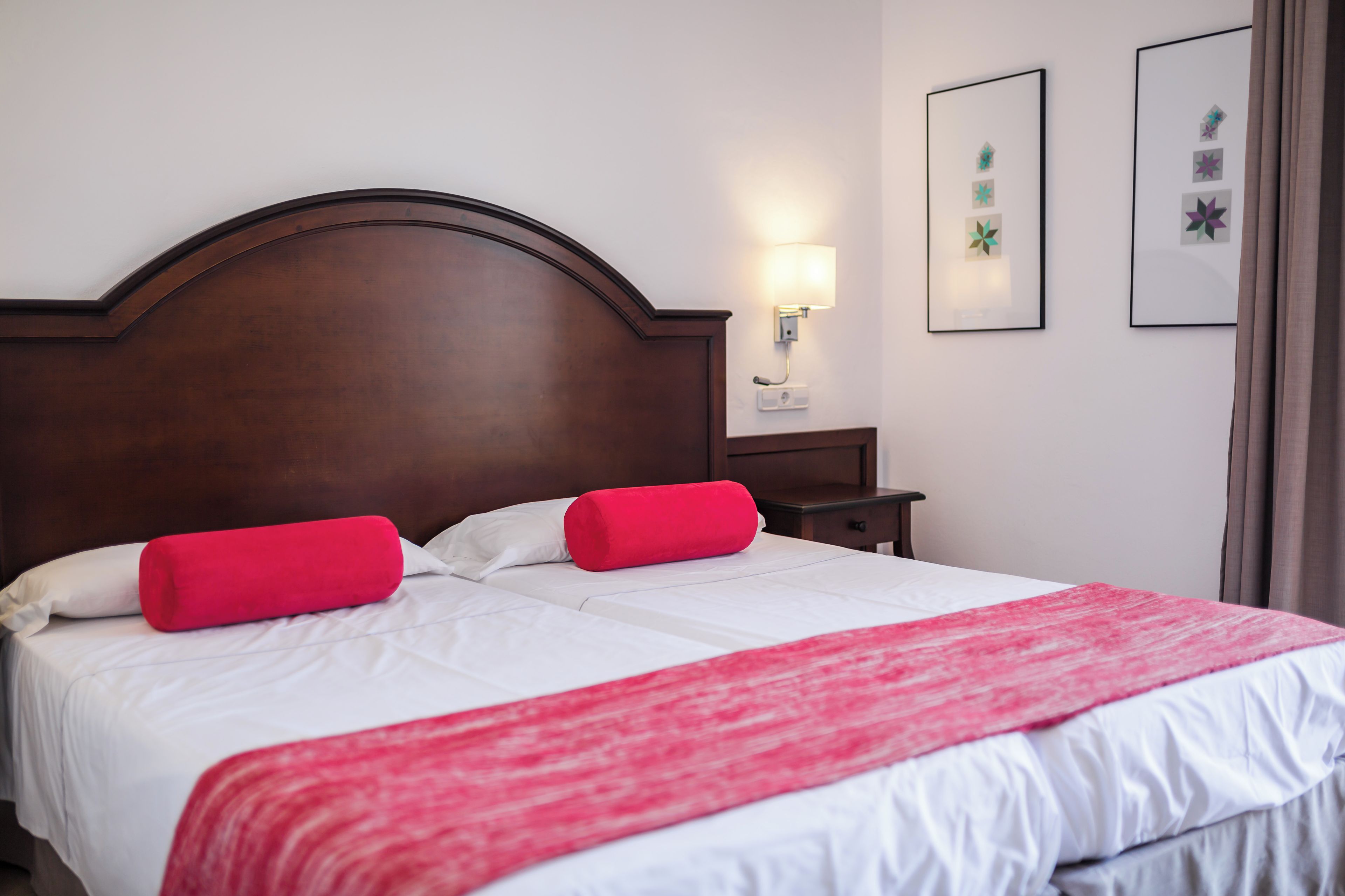 Una cama bien hecha con sábanas blancas y detalles en rojo, un cabecero de madera, una mesita de noche con lámpara y cuadros en la pared.