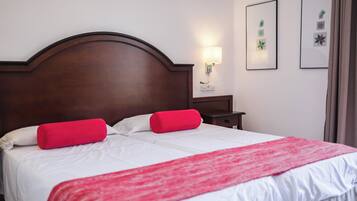 Una cama bien hecha con sábanas blancas y detalles en rojo, un cabecero de madera, una mesita de noche con lámpara y cuadros en la pared.