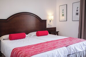 Un letto ben rifatto con lenzuola bianche e dettagli rossi, una testiera in legno, un comodino con una lampada e opere d'arte incorniciate appese al muro.