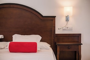 A neatly made bed with white and red pillows, a wooden headboard, a bedside table with a drawer, and a wall-mounted light fixture. - Ca'l Bisbe (Sóller)