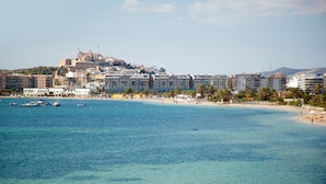 A coastal cityscape with a prominent hill, clear blue waters, and a beach with people and boats. - Hotel Ses Figueres (Ibiza Town)