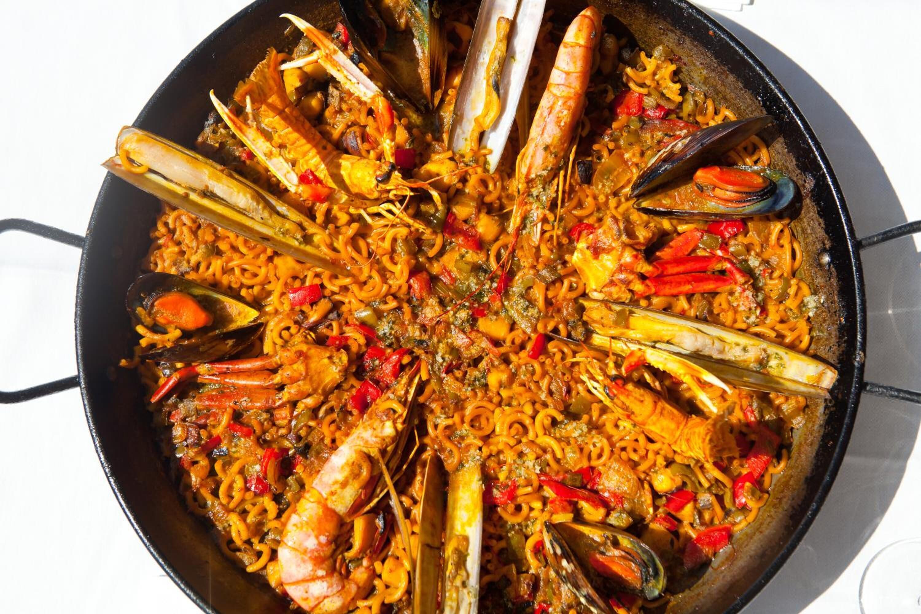 En stor, rund, svart stekpanna fylld med en paella med skaldjur, inklusive räkor, musslor och röda paprikor.