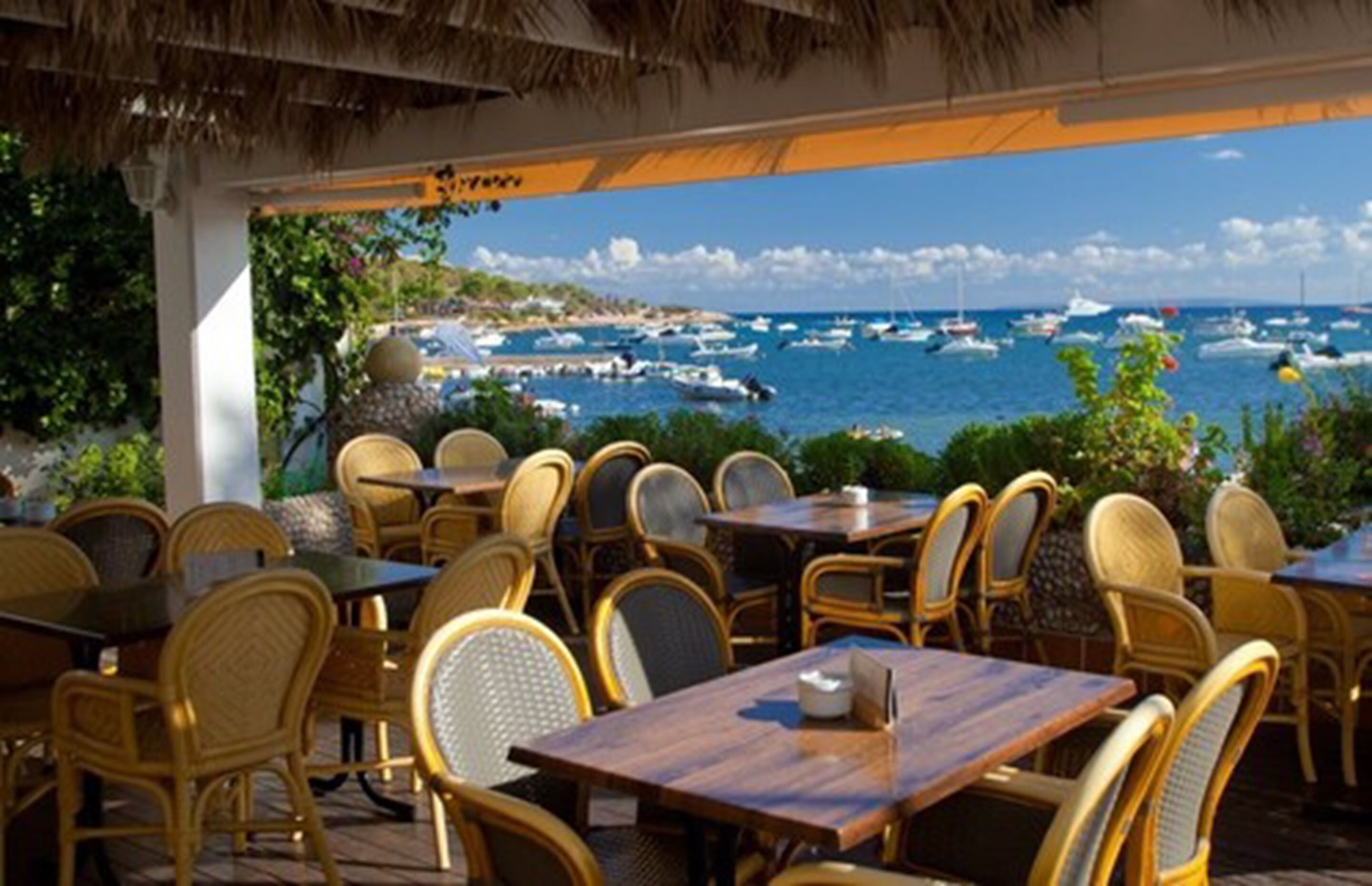 An outdoor seating area with wicker chairs and tables, overlooking a marina with boats.
