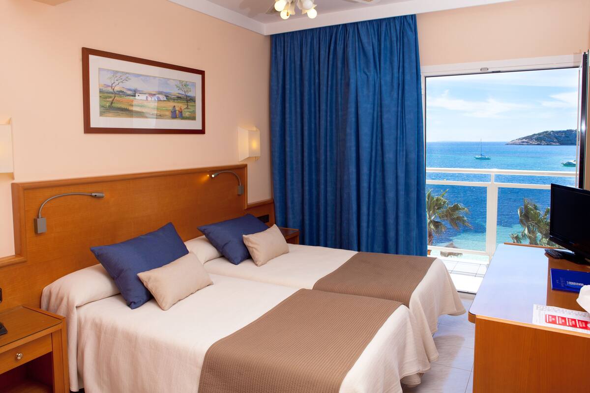 Una camera d'albergo con due letti, una scrivania, una televisione, un quadro appeso al muro e vista sul mare.