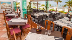 A restaurant with multiple tables and chairs, some with white tablecloths and starfish designs. There are bottles of beverages on the tables, and the restaurant overlooks a beach with palm trees and boats. - Hotel Ses Figueres (Ibiza Town)