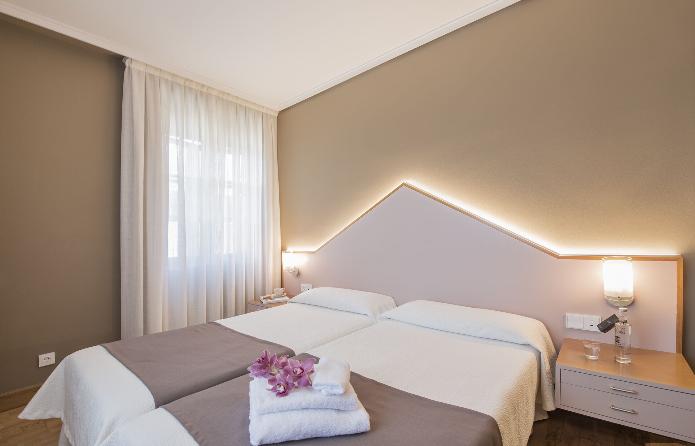 A hotel room with two beds, a nightstand with a lamp, and a window with curtains.