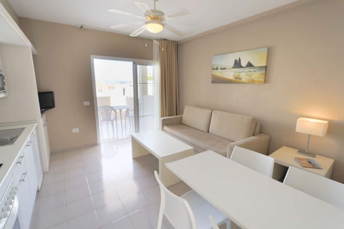 A modern hotel room with a kitchenette, a sofa, a dining table, and a painting on the wall.