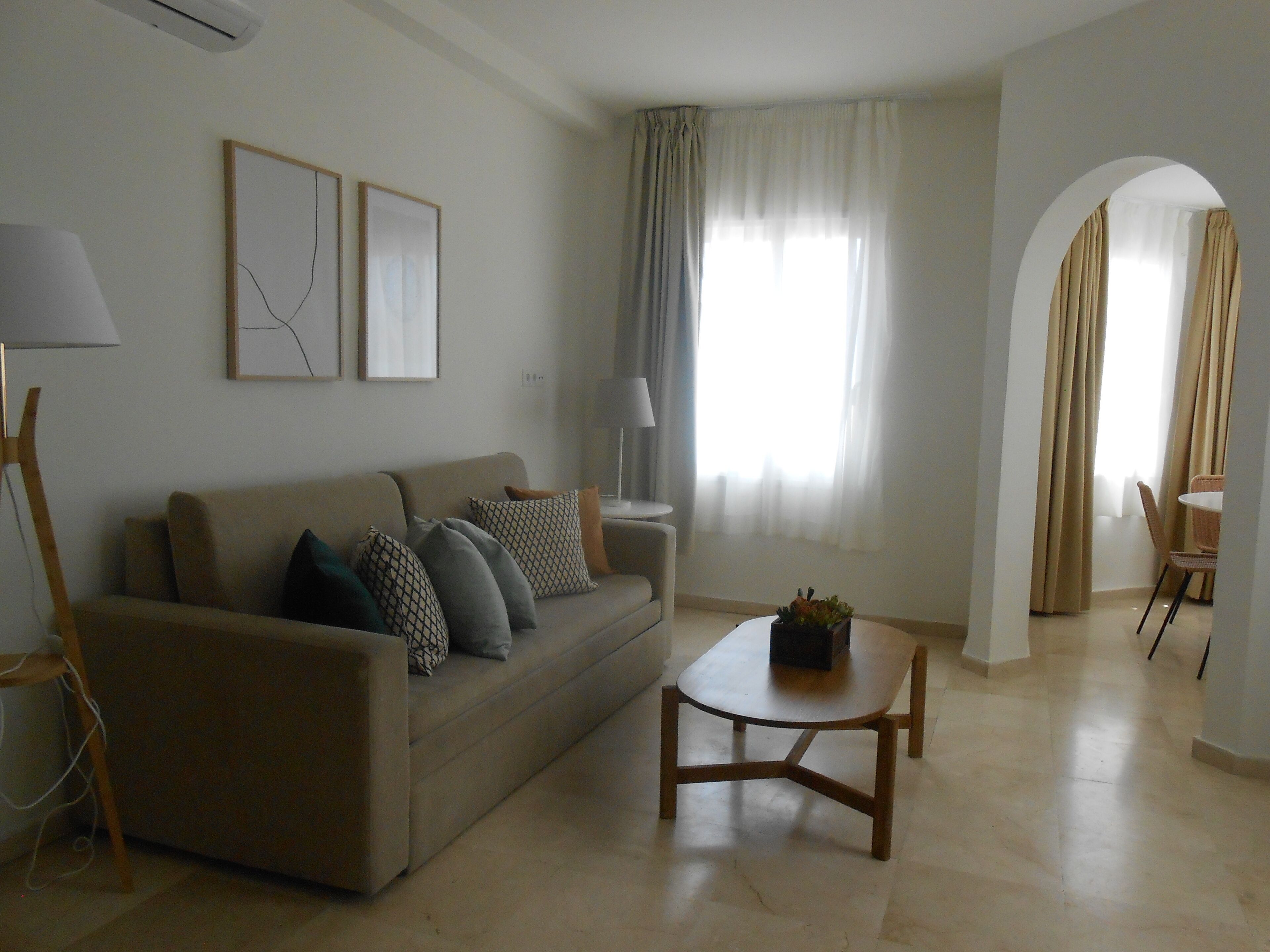 A living room with a sofa, a coffee table, and a lamp. There is a window with curtains and a door with a curtain.