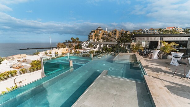 Een dakzwembad met oneindige rand gevuld met helderblauw water, met uitzicht op de oceaan en een resort in de verte.