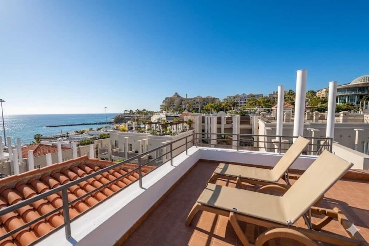 A rooftop terrace with lounge chairs, a pool, and a view of the sea and buildings.