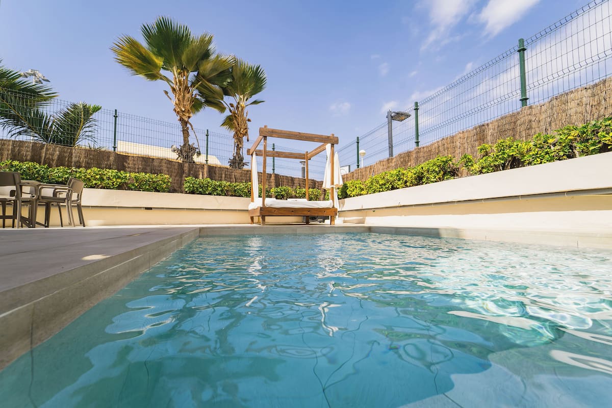 A swimming pool with a wooden structure, surrounded by a fence and palm trees.