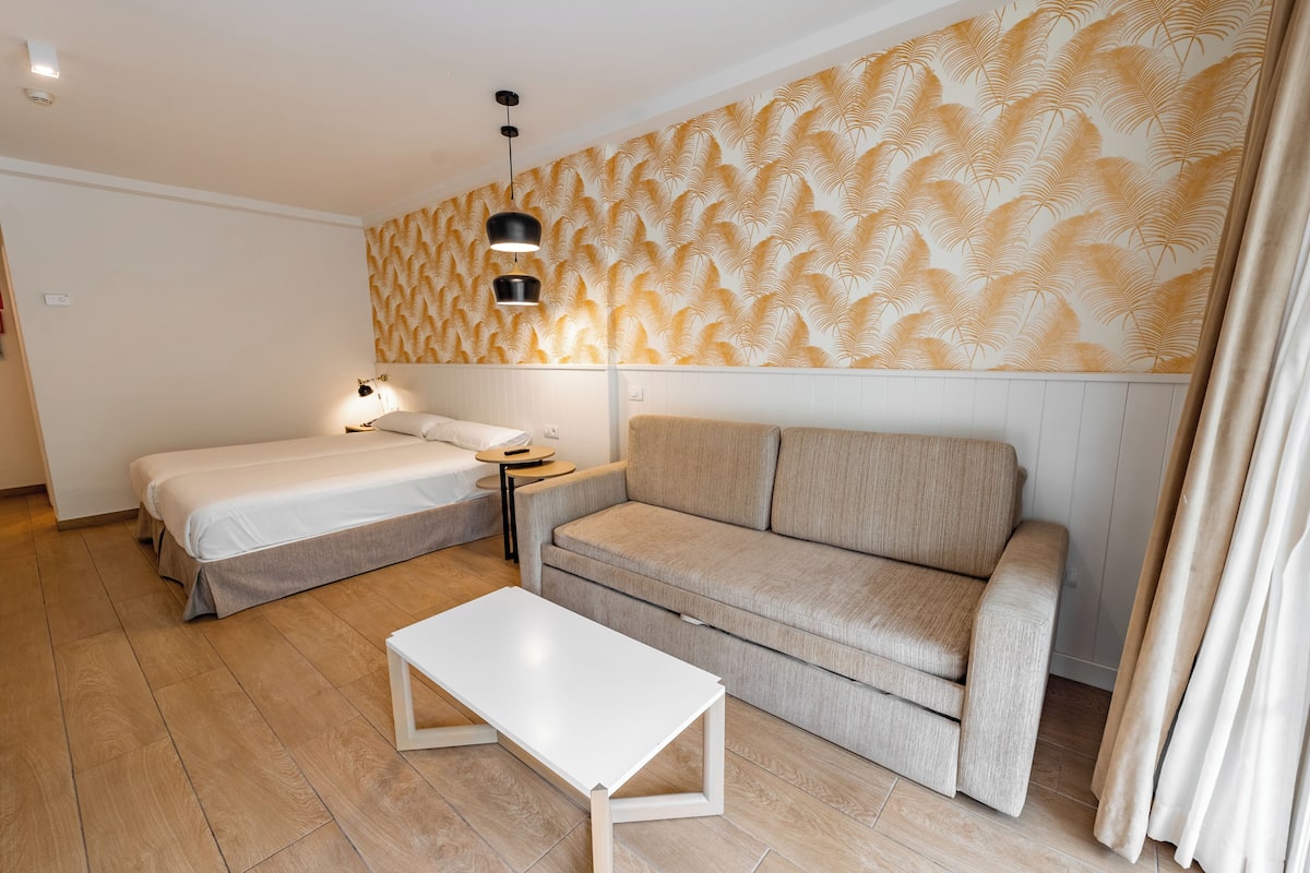 A room with two beds, a sofa, a white table, and patterned wallpaper.