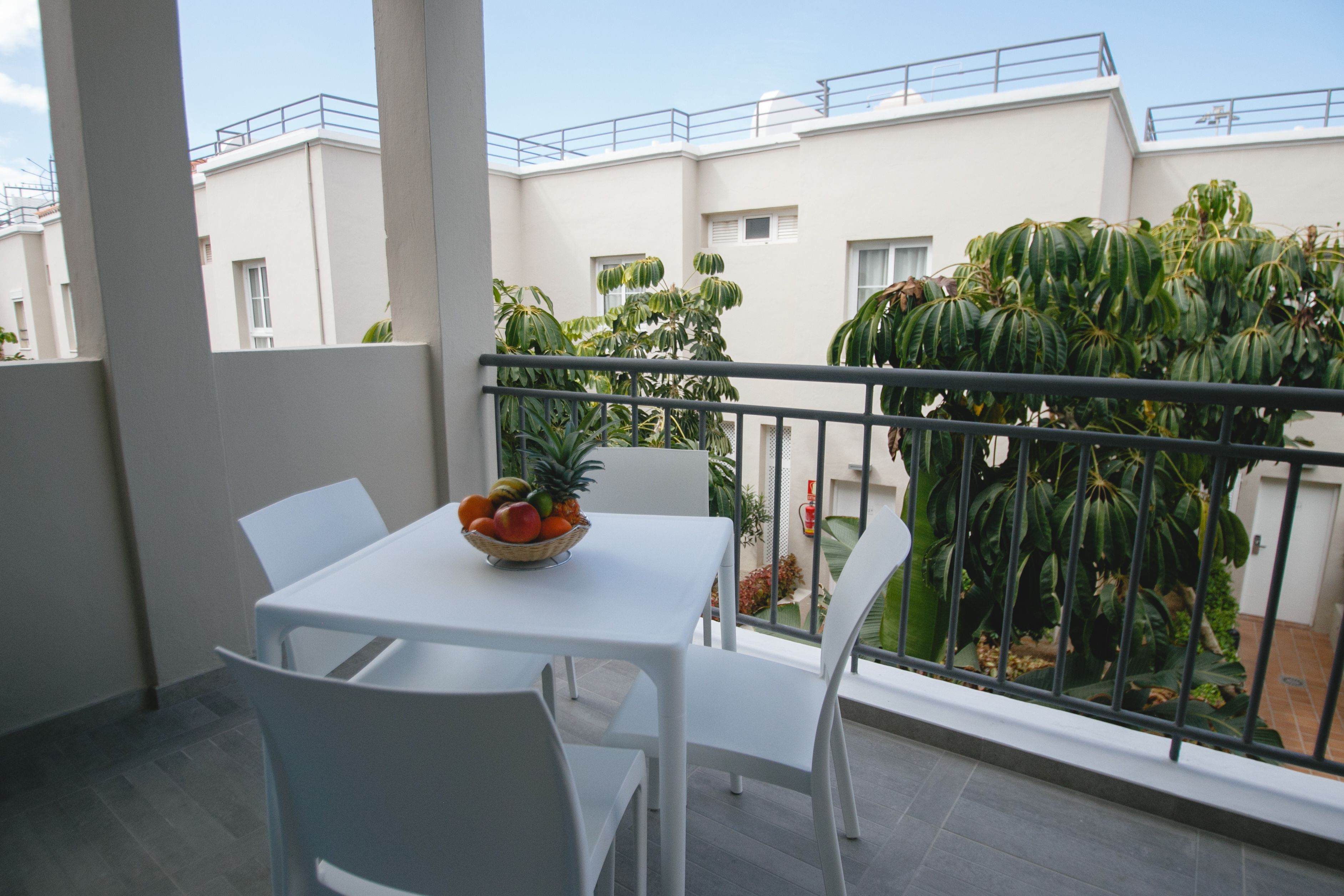 A balcony with a white table and chairs, a fruit bowl, and a view of a building and greenery.