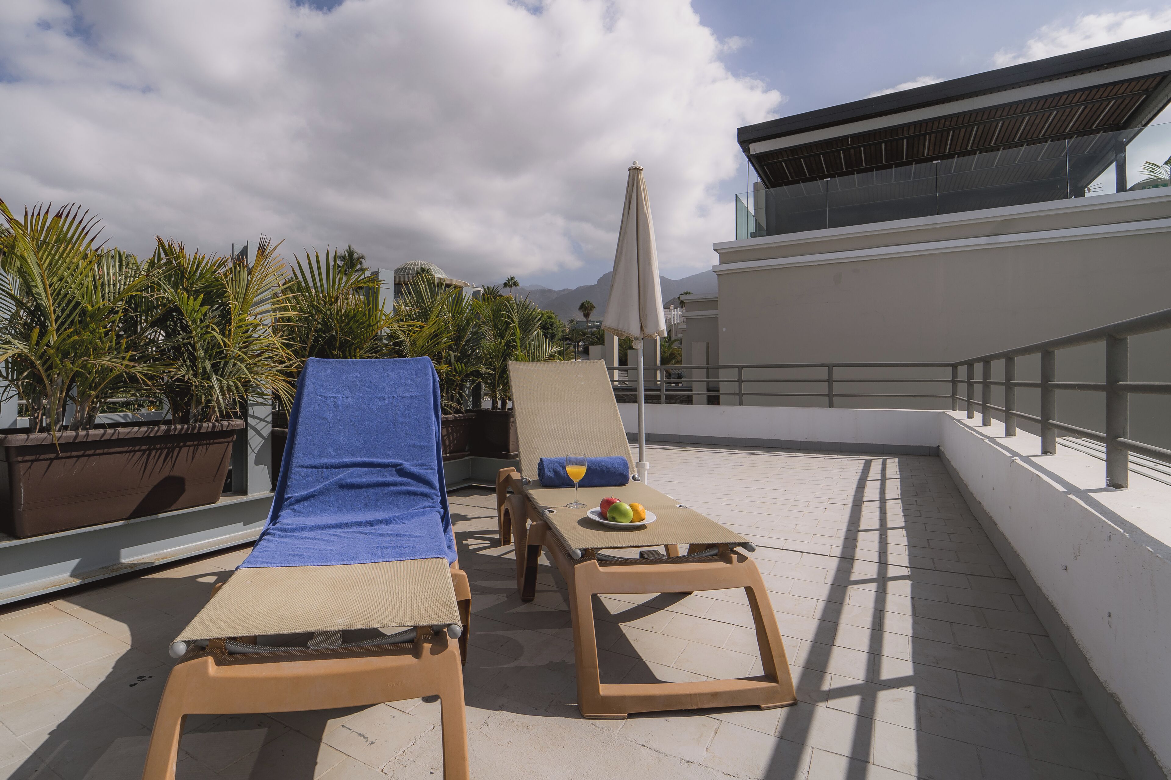 A rooftop terrace with two sun loungers, a table with fruit and a drink, and a large umbrella.
