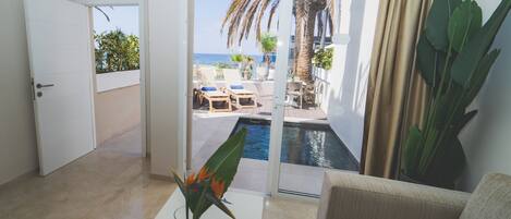 Quarto com vista para uma piscina e o mar, um sofá, uma mesa de centro com uma planta e uma porta que dá para uma área externa.