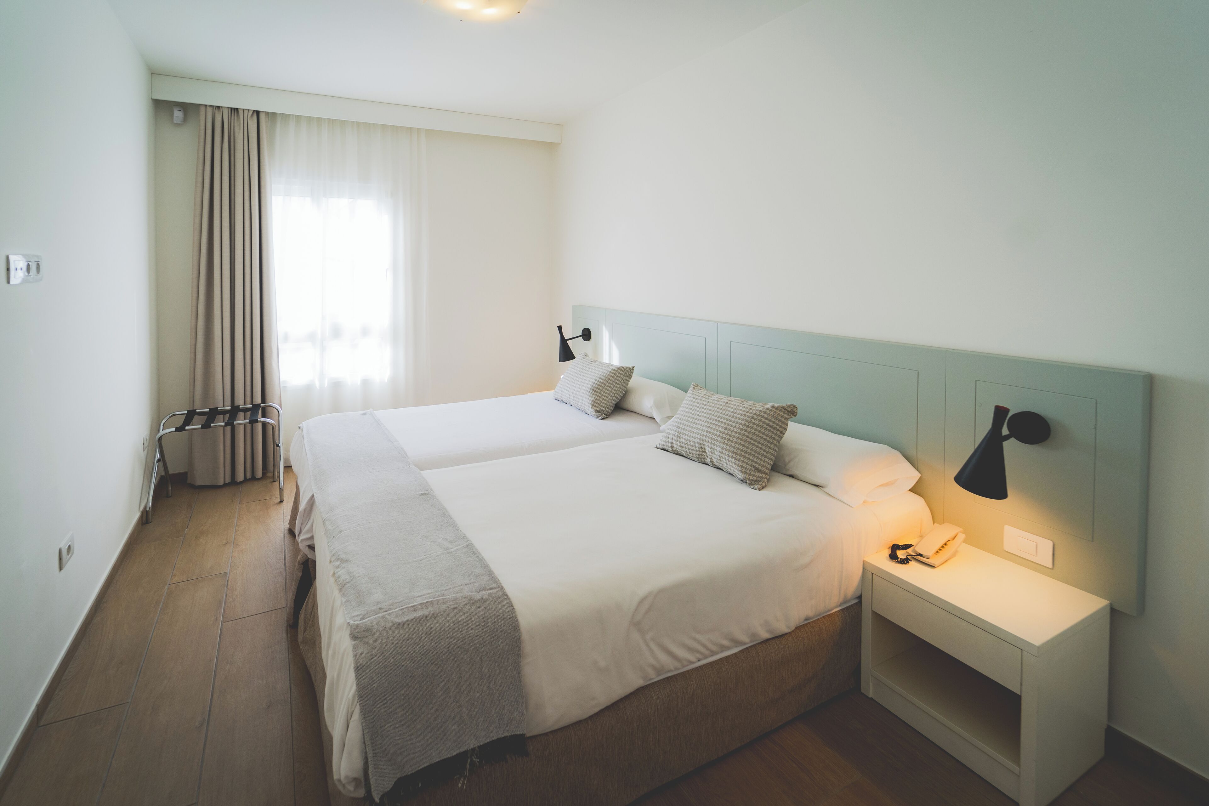 A hotel room with two beds, a nightstand with a lamp, and a window with curtains.