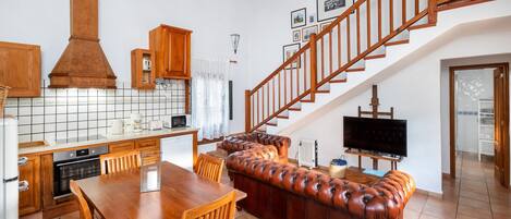 A spacious kitchen with wooden cabinets, a dining table, and a large sofa. A staircase leads to an upper level with a TV and framed pictures.